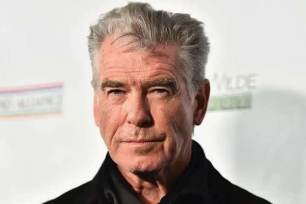 Portrait de Pierce Brosnan aux cheveux gris, en manteau noir, devant un fond clair