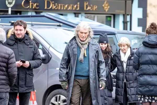 Pierce Brosnan aux cheveux longs marchant dans une rue animée devant un pub irlandais