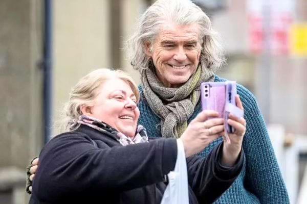 Pierce Brosnan aux cheveux gris posant pour un selfie avec une femme souriante