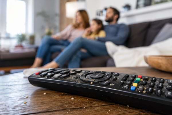 Télécommande sale pleine de miettes sur une table basse, devant une famille qui regarde la télévision