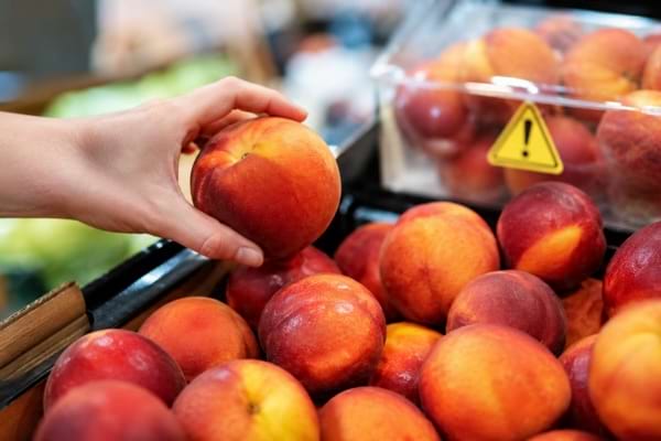 Main tenant une nectarine près d’une barquette signalée avec un pictogramme d’alerte.