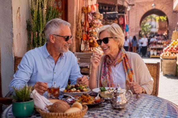 Couple de retraités souriant à table dans un café marocain près d’un marché coloré.