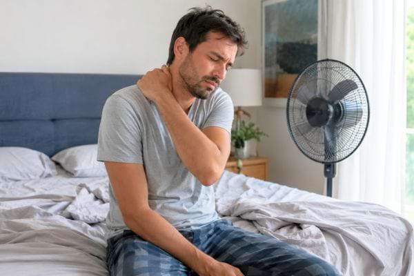 Homme assis sur son lit au r&eacute;veil, tenant sa nuque douloureuse pr&egrave;s d&rsquo;un ventilateur.