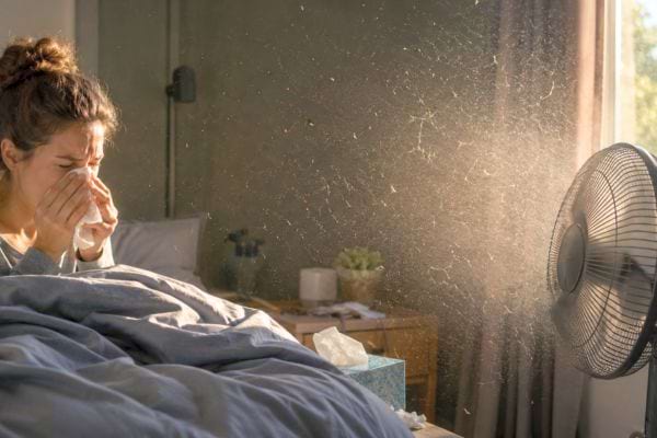 Femme au lit qui &eacute;ternue pendant qu&rsquo;un ventilateur brasse poussi&egrave;re et particules dans l&rsquo;air.