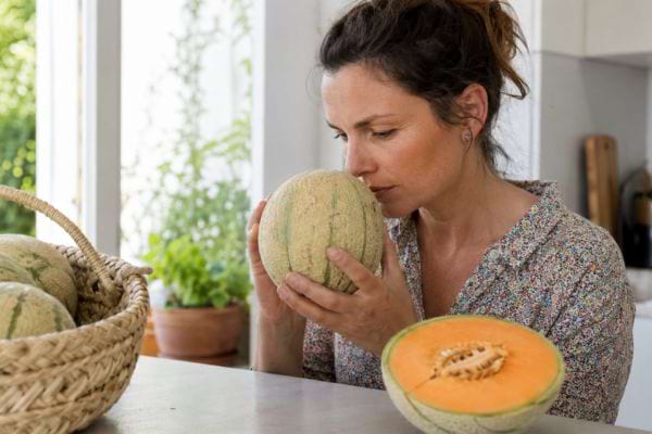 Femme sentant un melon entier dans sa cuisine, près d’un melon ouvert à la chair orangée.