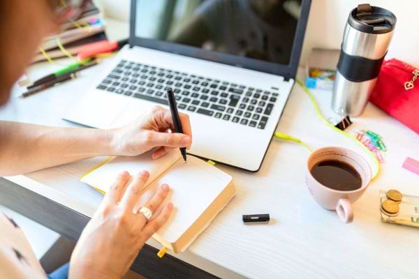 gaucher qui écrit avec la main gauche sur un carnet devant son ordinateur