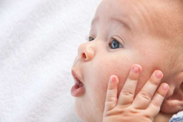 un bébé aux yeux bleu fait une tête étonnée et pose sa main sur la joue