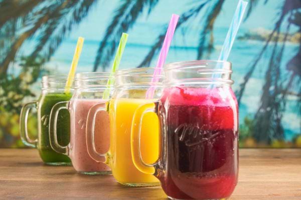 4 verres de jus de fruits colorés avec des pailles devant un paysage de mer et cocotoer