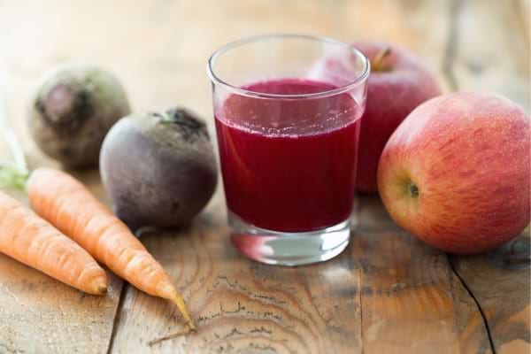 Jus de betterave, pomme et carotte servi dans un verre sur une table en bois.