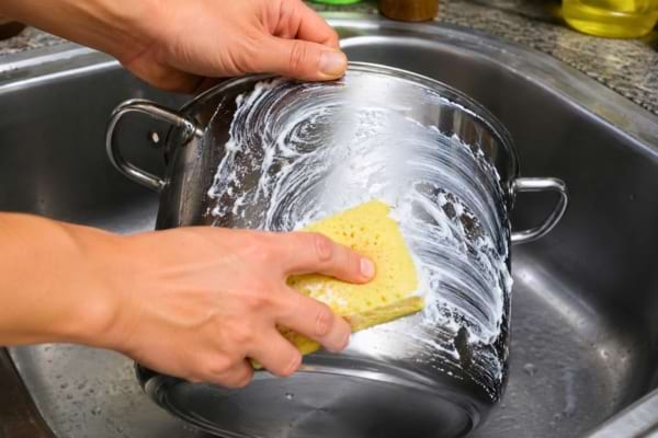 A shiny stainless steel cooking pot in a kitchen sink, being gently scrubbed with a thick white paste and a yellow sponge