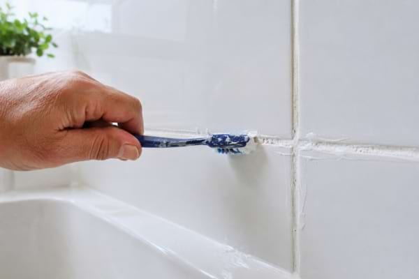 A person's hand using an old toothbrush to scrub white bathroom tile grout with a thick white cleaning paste