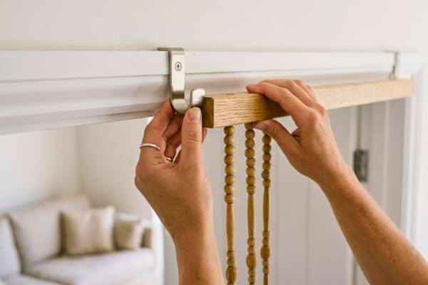 mains fixant d&eacute;licatement une tringle en bois avec des perles sur deux crochets m&eacute;talliques
