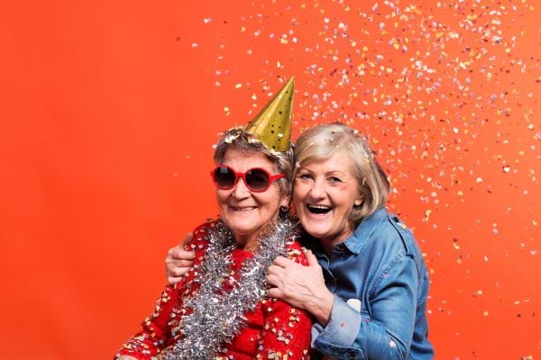 deux femmes âgées qui font la fête avec des confettis et des chapeaux sur un fond orange
