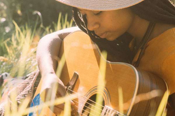 une jeune femme noire style hippie qui joue de la guitare dans un champs un chapeau sur la tête