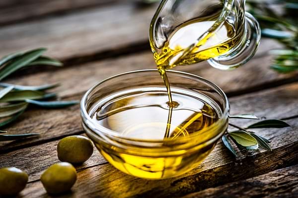 Huile d’olive versée dans un bol en verre entouré d’olives vertes.