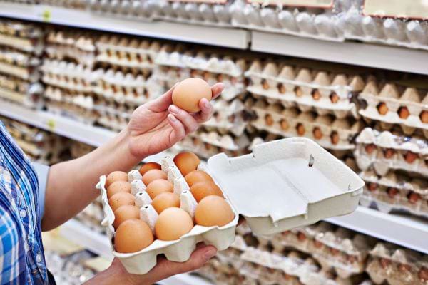une personne tient un oeuf dans la main devant un rayon d'oeufs au supermarch&eacute;