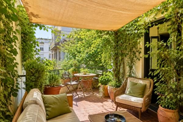 a lush, green apartment balcony in summer. A light beige shade sail is stretched overhead, blocking the harsh sun