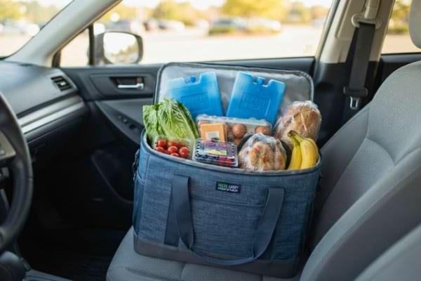A reusable insulated cooler bag with ice packs and fresh groceries inside, sitting on a car seat