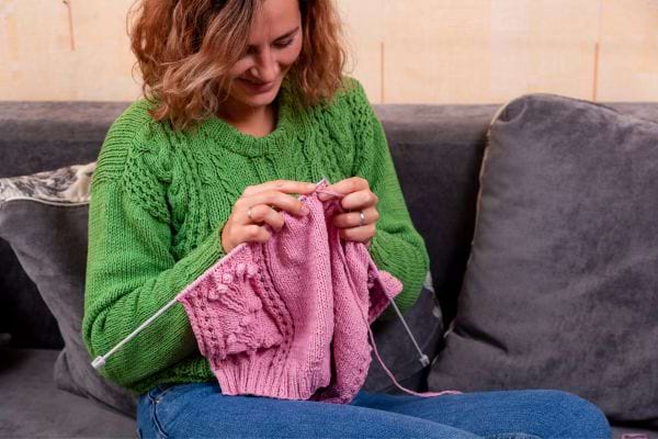 une femme souriante avec un pull vert en train de tricoter un pull rose
