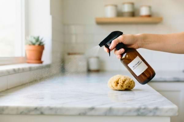 une main tenant un vaporisateur en verre ambr&eacute;, en train de vaporiser un plan de travail de cuisine en marbre