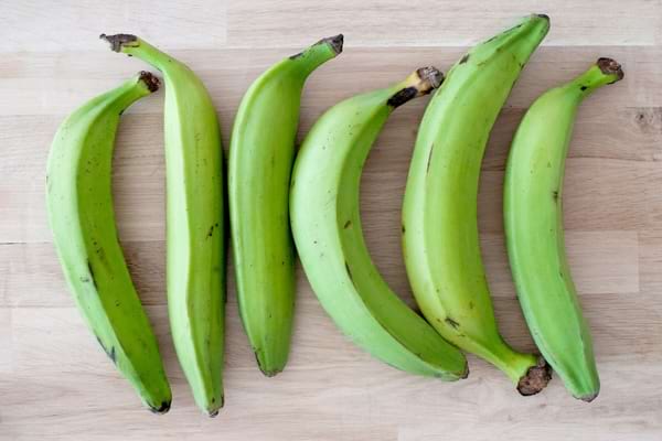 Six bananes plantains vertes alignées sur une table en bois.