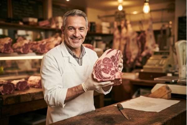 Un artisan boucher souriant en tablier blanc, se tenant derrière son comptoir en bois, présentant une belle pièce de bœuf