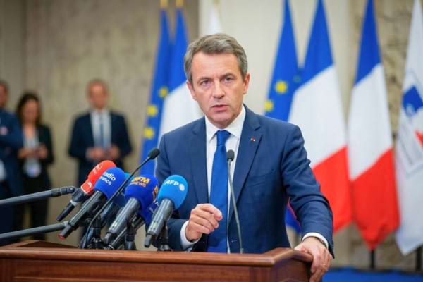 un homme politique en costume cravate, l'air sérieux et concentré, s'exprimant derrière des micros