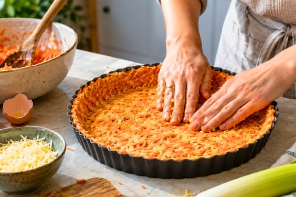 Mains qui tassent un mélange de carottes râpées, d'œufs et de fromage dans un moule à tarte.