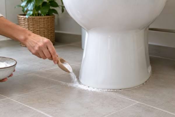 Main saupoudrant du bicarbonate de soude autour du pied des toilettes.